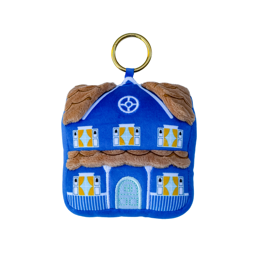 A blue felt house-shaped keychain or bag charm with a gold metal ring, featuring brown faux-fur trim, yellow window shutters, and a light blue front door on a white background.
