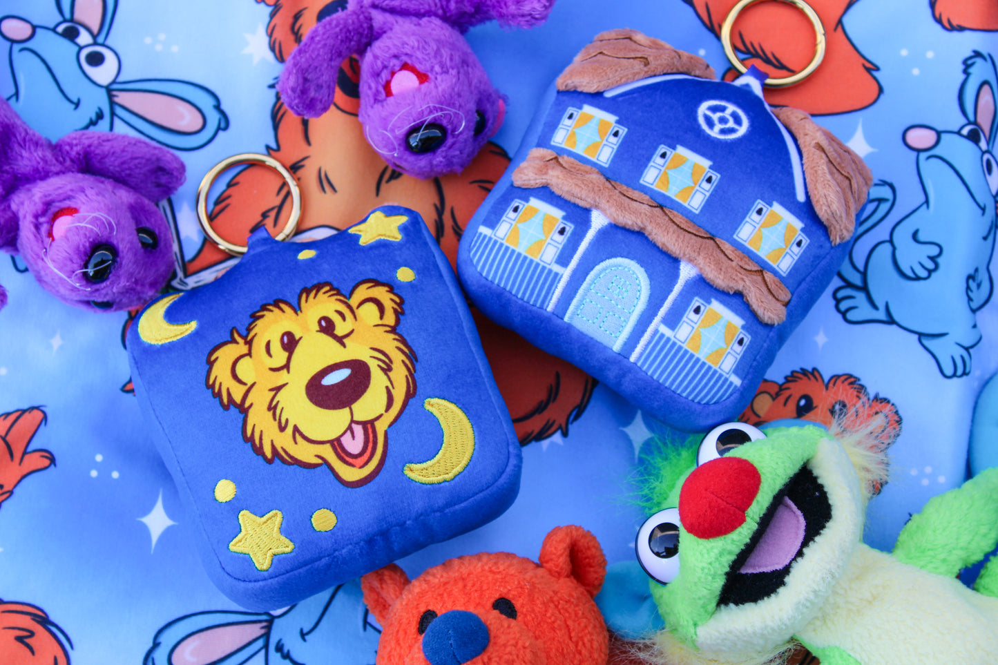 A close-up flat lay of plush character accessories on a blue printed fabric background. The main products are two square plush pouches or cases: one blue with a yellow lion face and moon/star embroidery, and one blue with a house design.