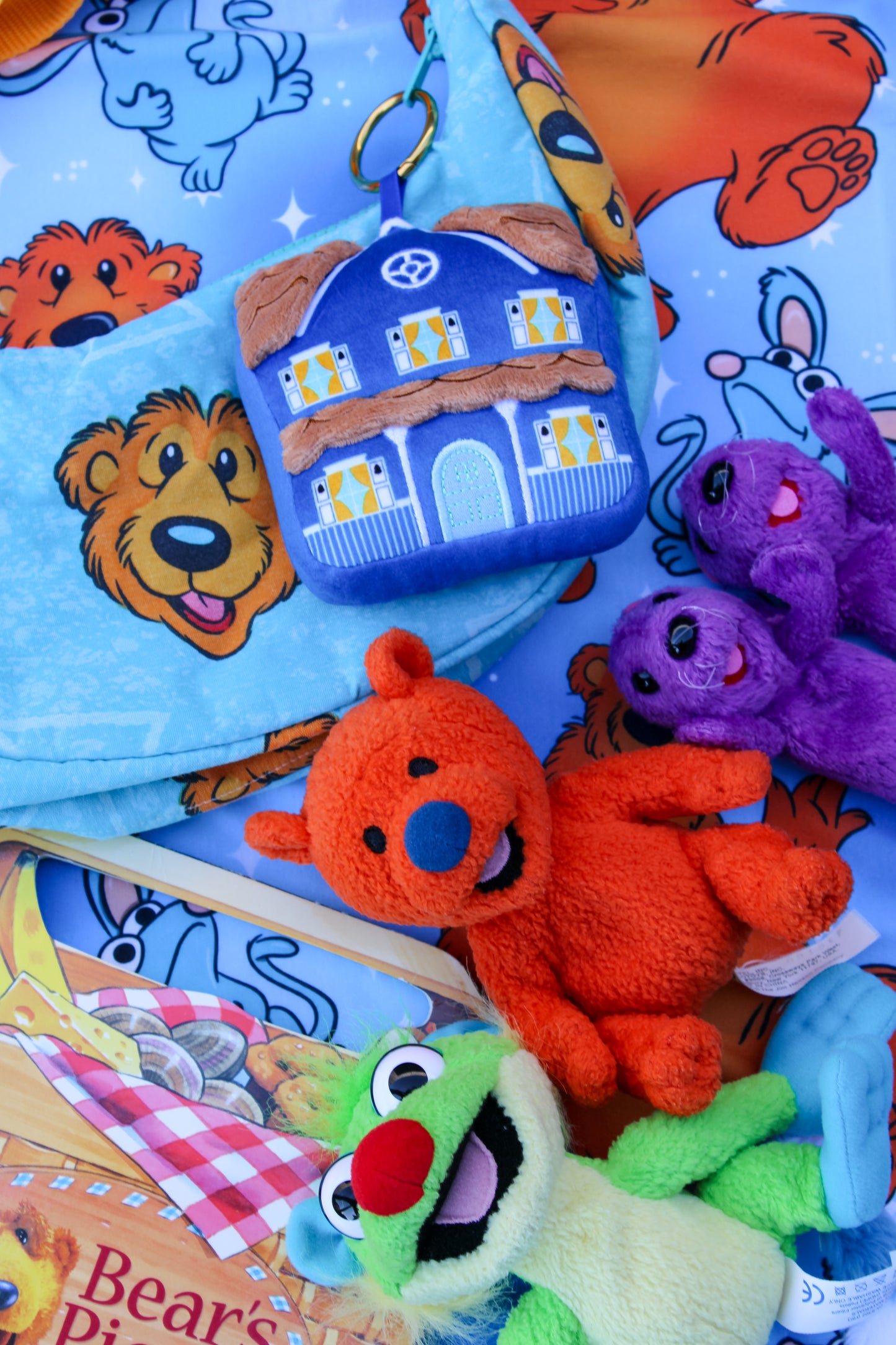 A collection of Muppet-themed plush toys and accessories, including a green Kermit the Frog plush, an orange Bear Bunchie plush, a purple Miss Piggy plush, and a blue house-shaped Airplay case, all arranged on a blue patterned blanket featuring cartoon bear faces.